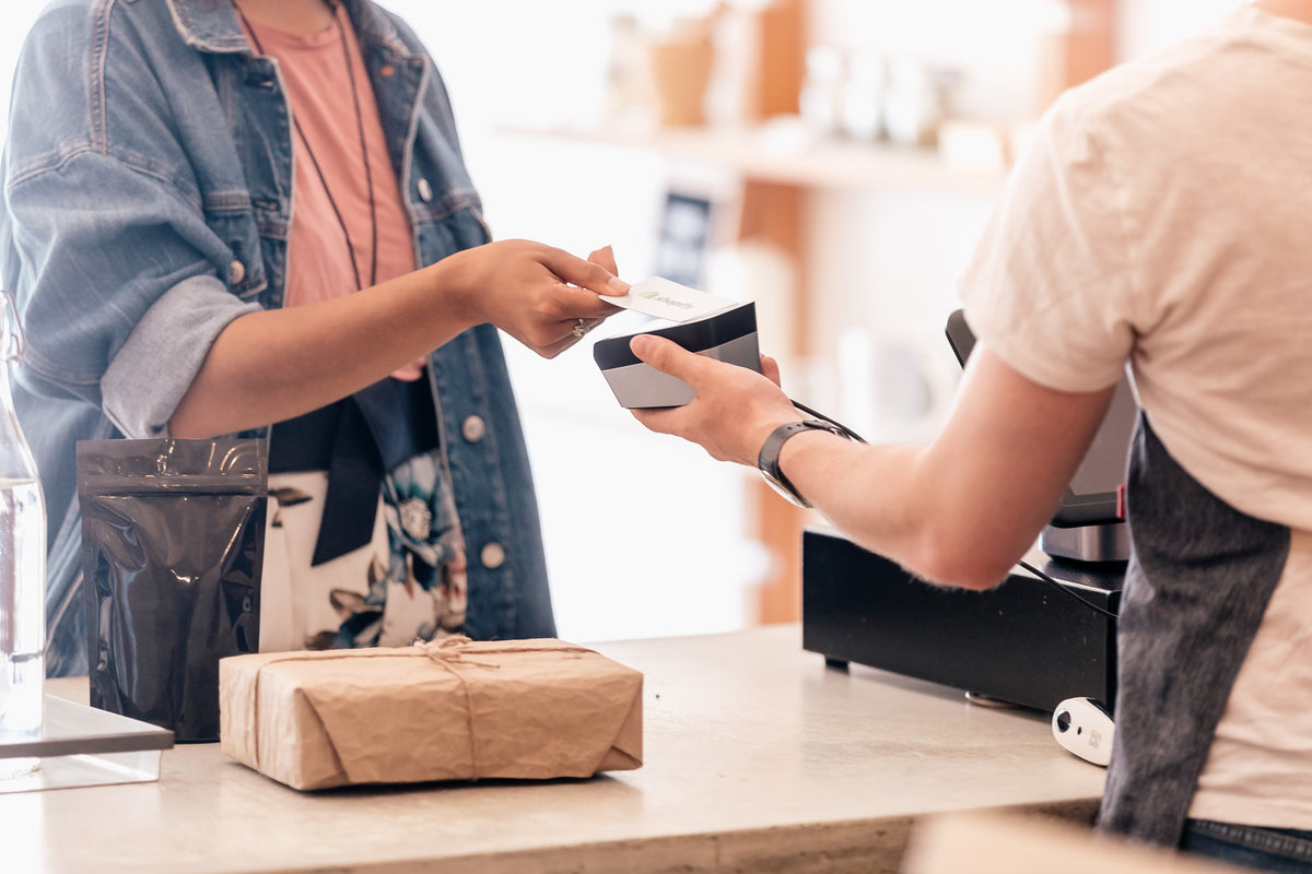 Eine Frau  bezahlt einkäufe mit ihrer Kreditkarte. Ein Päckchen, das in naturbelassenes Papier eingepackt ist, liegt auf dem Verkaufstresen.